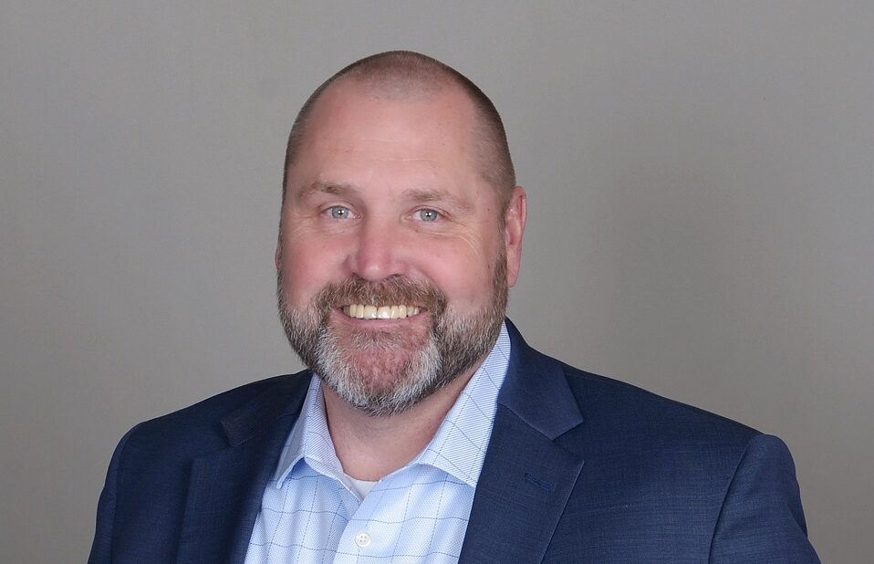 Portrait of a smiling man with a beard, wearing a navy blazer and light blue checked shirt against a gray background.